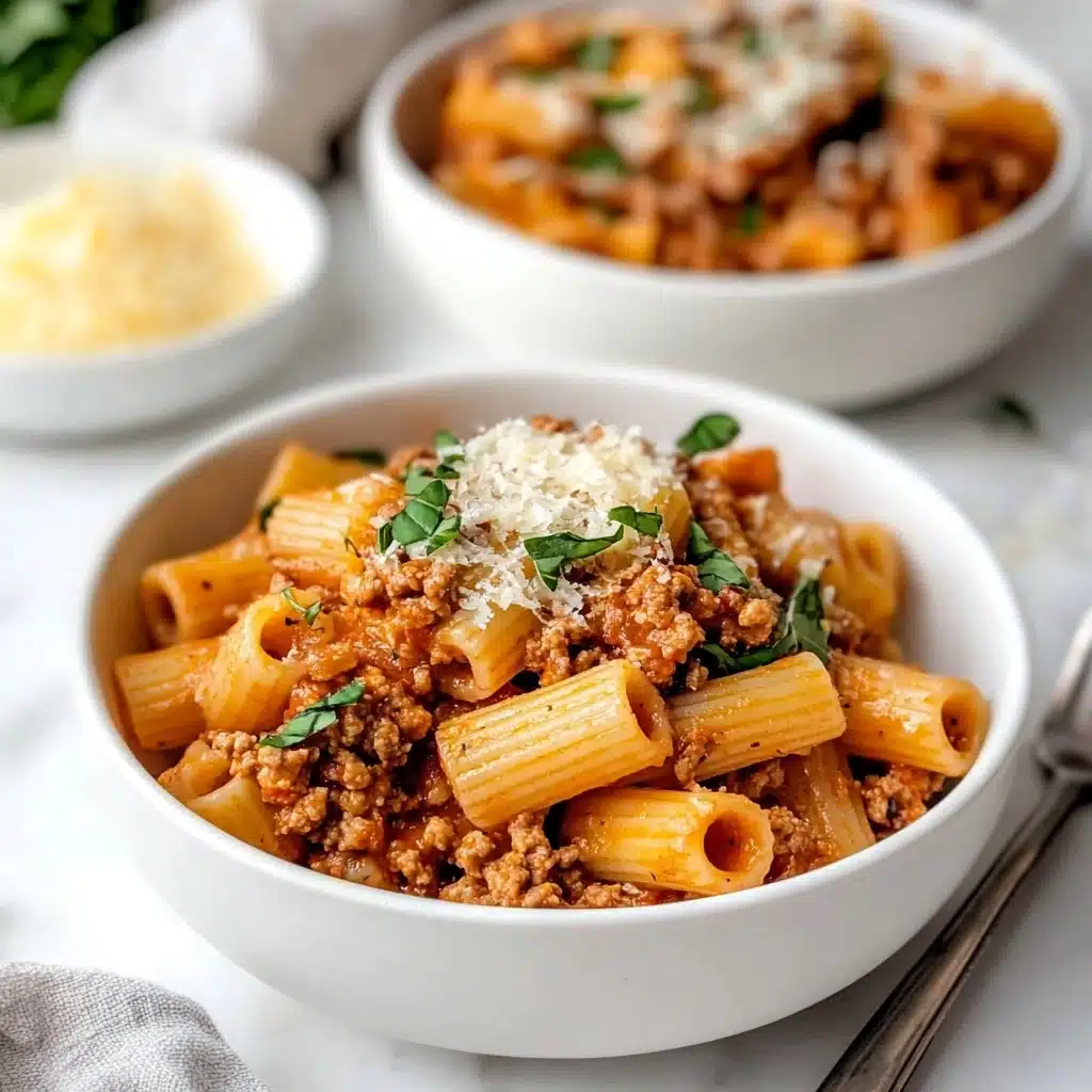 Rigatoni with Spicy Sausage and Creamy Tomato Sauce, spicy sausage pasta, creamy tomato sauce recipes, easy Italian pasta dishes, hearty sausage pasta meal Two white bowls on a white marbled surface each hold a serving of rigatoni pasta. The pasta is coated in a thick, reddish-brown meat sauce with small pieces of ground meat mixed in. The dish is topped with green leafy herbs and sprinkled with grated cheese. In the background, there is a small bowl with extra grated cheese and a light gray cloth. A silver fork rests on the white marbled surface beside the nearest bowl.