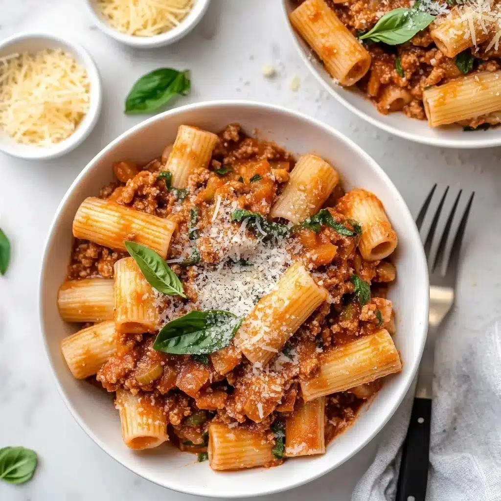 Rigatoni with Spicy Sausage and Creamy Tomato Sauce, spicy sausage pasta, creamy tomato sauce recipes, easy Italian pasta dishes, hearty sausage pasta meal The image shows two white bowls filled with rigatoni pasta layered with a thick, creamy reddish-brown sauce mixed with ground meat and small bits of vegetables. The pasta tubes are large and arranged to show texture on their surface, sitting in a generous layer of sauce. On top is a sprinkle of finely grated light-colored cheese and fresh green basil leaves scattered for color contrast. Surrounding the bowls are small white ramekins, one filled with grated cheese and another with fresh chopped green herbs. The scene rests on a clean white marbled surface with a metal fork featuring a dark handle placed near the nearest bowl. The lighting is soft and natural, making the food look fresh and inviting. Woman’s hand is not visible in this frame.