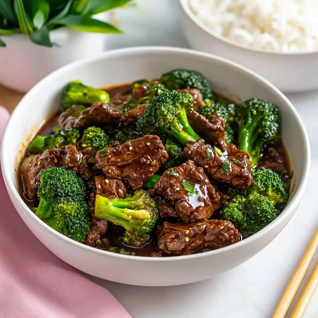 Chinese Beef and Broccoli, Chinese Beef and Broccoli stir-fry, homemade Chinese beef and broccoli, easy Chinese beef and broccoli recipe, restaurant-style beef and broccoli A white bowl filled with beef and broccoli stir fry placed on a white marbled surface with a pink napkin in the front. The dish has two main layers: bright green broccoli florets with visible water droplets and dark brown chunks of tender beef coated in a glossy brown sauce with small bits of garlic. In the background to the right, there is a white bowl filled with white rice. Part of a small green plant in a white pot is visible in the top left corner. A pair of chopsticks with pink tips lies on the bottom right. The