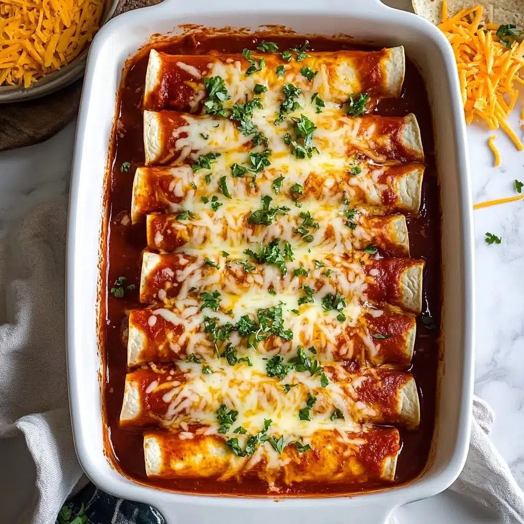 Homemade Easy Beef Enchiladas, beef enchiladas, homemade enchiladas, easy enchilada recipe, Mexican beef enchiladas A white rectangular dish holds eight rolled tortillas placed side by side in two rows, each tortilla lightly browned on the edges. They are covered with a thick red sauce, mostly covering the tortillas but leaving some white edges visible. A layer of melted orange and yellow shredded cheese covers the center of the tortillas, topped with scattered green chopped herbs for garnish. The dish is on a white marbled surface with some shredded cheese and a bit of cloth visible near it.
