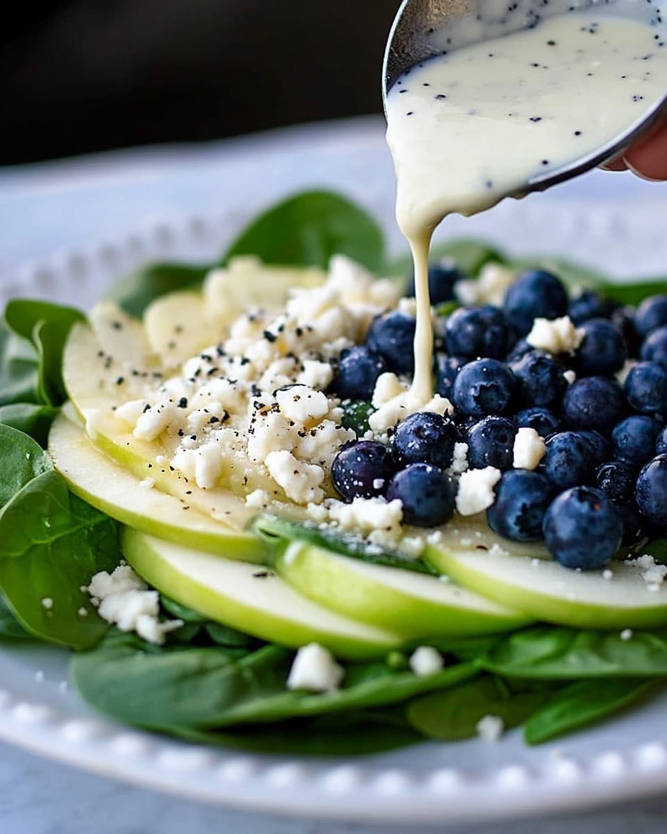 Blueberry Spinach Salad with Homemade Orange Poppy Seed Dressing, healthy blueberry spinach salad, fresh fruit and greens salad, easy homemade salad dressing, summer fruit salad A white plate holds a salad made of three layers: the bottom layer is fresh dark green spinach leaves, the middle layer is thin slices of light green apple with small white nuts scattered around, and the top layer is a pile of dark blue blueberries. White cheese crumbles are lightly spread on the salad. Above the plate, a woman's hand is holding a spoon pouring creamy white dressing with small black specks onto the salad, and the plate is set on a white marbled surface.