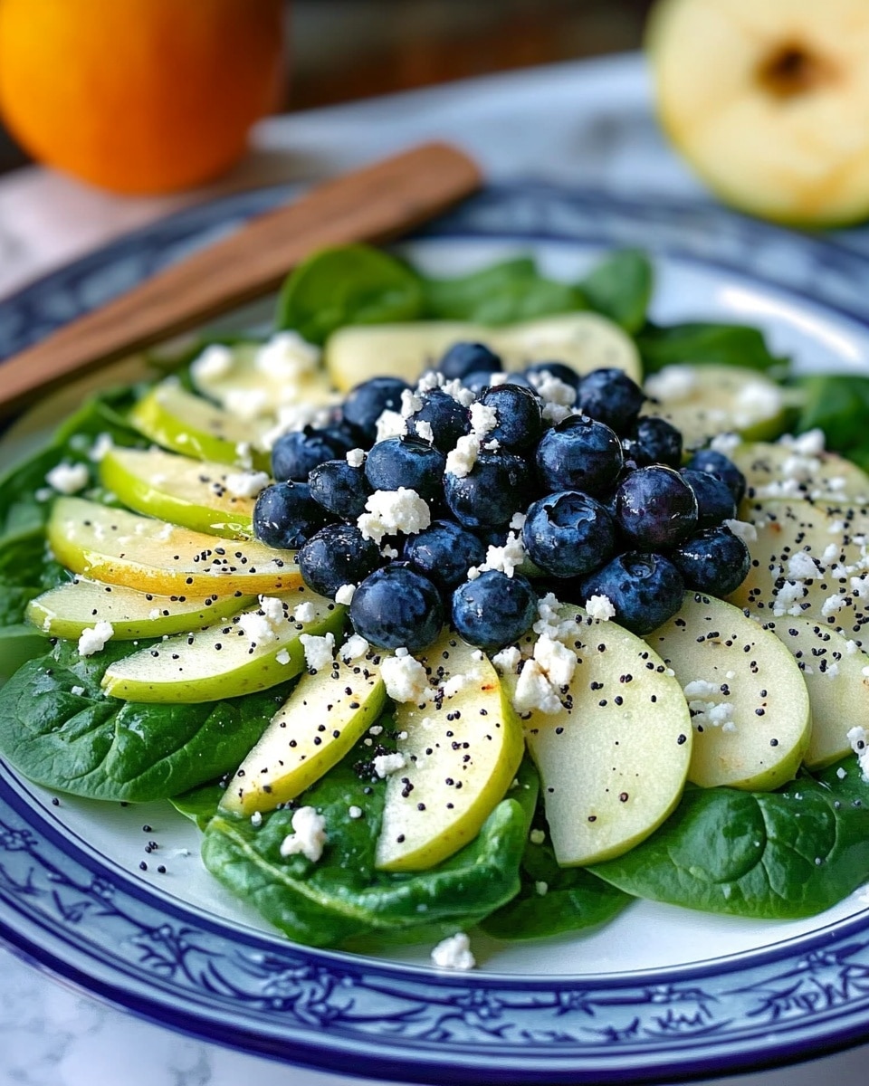 Blueberry Spinach Salad with Homemade Orange Poppy Seed Dressing, healthy blueberry spinach salad, fresh fruit and greens salad, easy homemade salad dressing, summer fruit salad A close-up view of a fresh salad on a white plate, resting on a white marbled surface. The salad has a base of green leafy spinach covering the whole plate. On top, there are thin slices of pale yellow cheese evenly placed in a circular layer. In the center, a cluster of small, round, dark blue blueberries adds contrast. To the side, small bits of white nuts are sprinkled over the greens. A woman's hand holding a silver fork is about to pick up the salad.