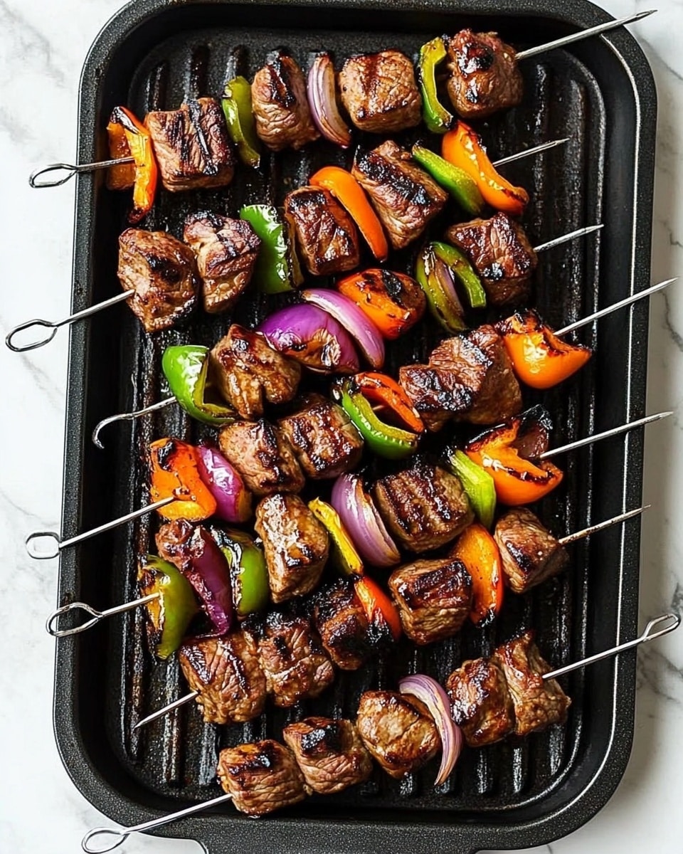 Beef Shish Kabob, Beef Shish Kabob Recipe, grilled beef skewers, easy shish kabob marinade, vegetable shish kebabs The image shows four metal skewers laid out neatly on a white tray with a slightly raised edge. Each skewer has alternating pieces of dark brown grilled meat chunks and colorful grilled vegetables, including slices of bright orange, green, and yellow bell peppers, and purple onion wedges. The meat pieces have a slightly charred texture, and the vegetables show a light grill with a slightly softened look. Some green herbs are sprinkled lightly over the skewers. A woman's hand is visible on the side, reaching toward the tray. The tray is placed on a white marbled surface.