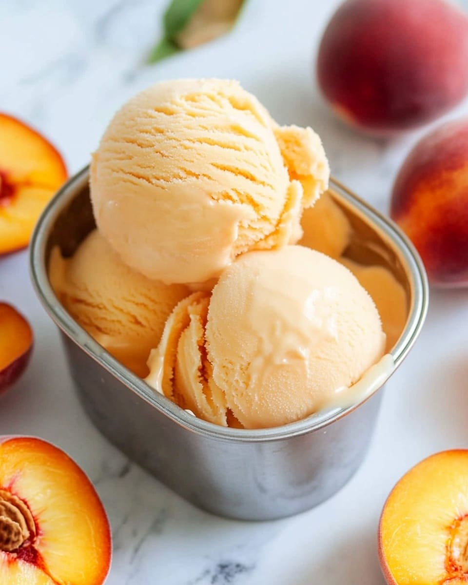 A small metal container filled with three large scoops of smooth, creamy peach-colored ice cream, showing soft ridges and slight melting around the edges. Around the container, there are whole and cut peaches with vibrant orange and red hues, resting on a white marbled surface. The metal container's silver color reflects some light, enhancing the fresh and cool look of the ice cream. Photo taken with an iphone --ar 4:5 --v 6.1