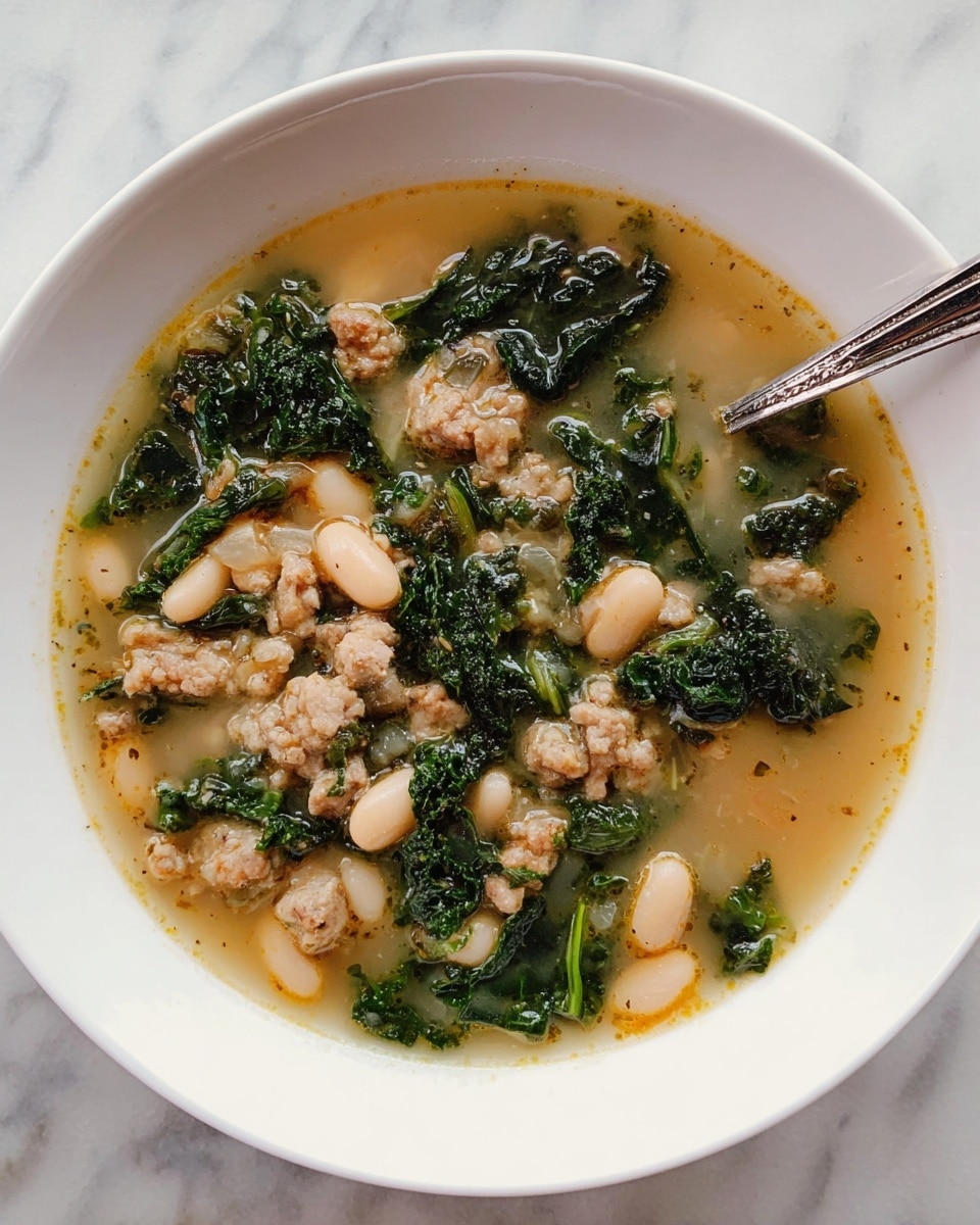 A white bowl filled with a soup that has three main layers: the bottom layer is a light brown broth with a slightly oily texture, the middle layer has white beans scattered within, and the top layer consists of dark green leafy kale pieces and small chunks of light brown cooked meat mixed evenly throughout. A silver spoon rests inside the bowl, partially submerged on the right side. The bowl sits on a white marbled surface. photo taken with an iphone --ar 4:5 --v 7