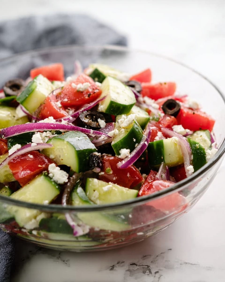 A clear glass bowl filled with a fresh salad made of chopped cucumber slices that are green with dark green skin, bright red tomato chunks, thin purple onion strips, small white feta cheese pieces, and black sliced olives scattered on top, all mixed together with visible specks of seasoning on a white marbled surface, with a blurred gray cloth in the background; photo taken with an iphone --ar 4:5 --v 7