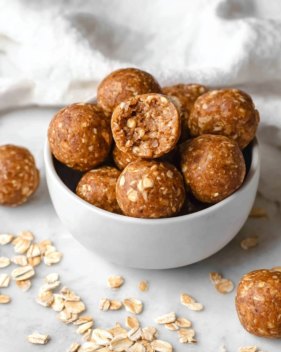 The image shows a white bowl filled with round energy bites that have a light brown color with oats visible inside. One energy bite is placed on top and is open, revealing a textured inside with small bits of oats and nuts. Around the bowl, there are similar energy bites scattered on a white marbled surface with a white cloth in the background. The overall look is warm and homemade. photo taken with an iphone --ar 4:5 --v 7