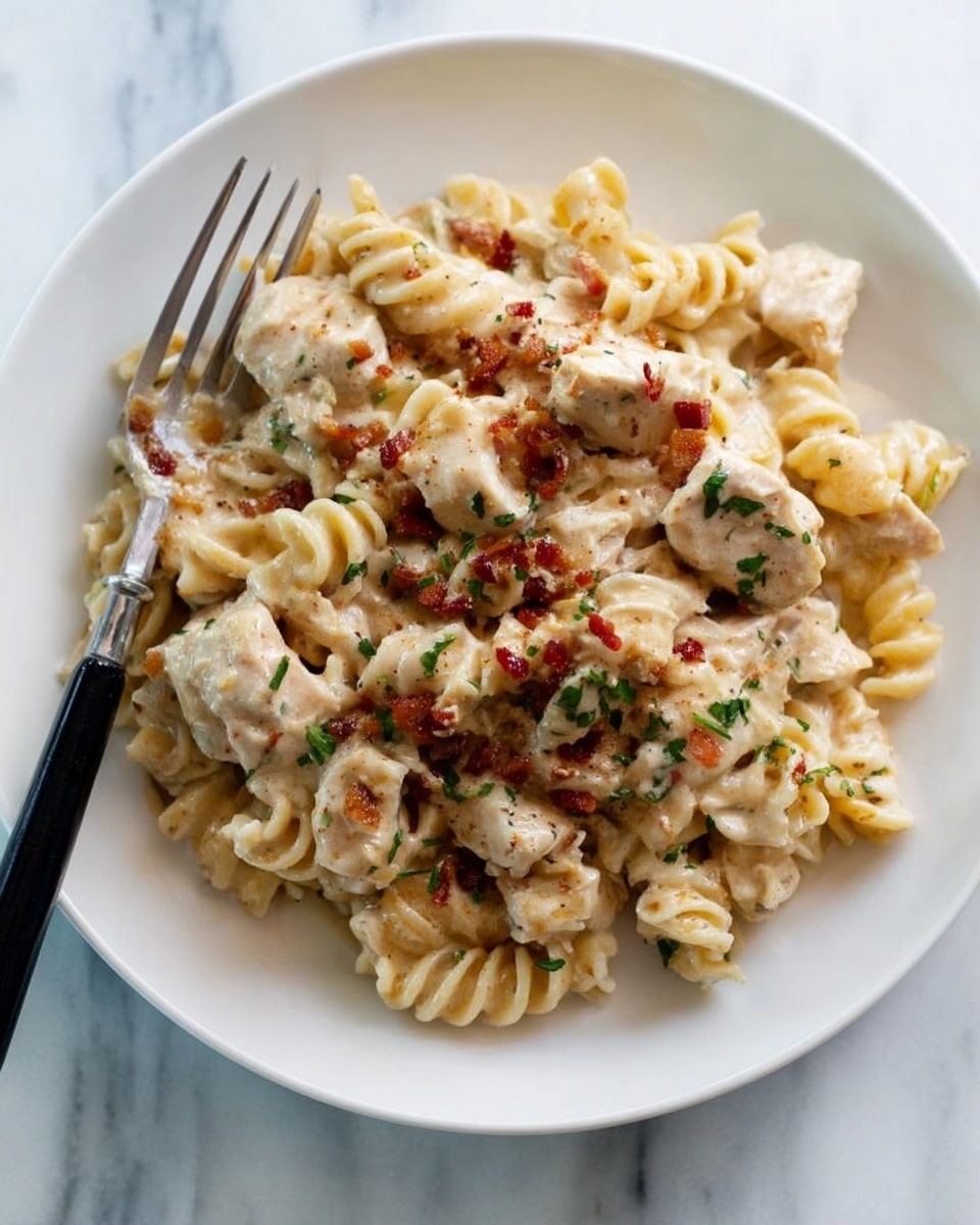 A white plate holds a creamy pasta dish with about three layers of short twisty pasta, covered in a light beige sauce with small red bits scattered on top. Some pieces of light brown chicken are mixed in and there are tiny green herb bits sprinkled through the pasta. A fork with a black handle rests on the edge of the plate. The plate sits on a white marbled surface. photo taken with an iphone --ar 4:5 --v 7
