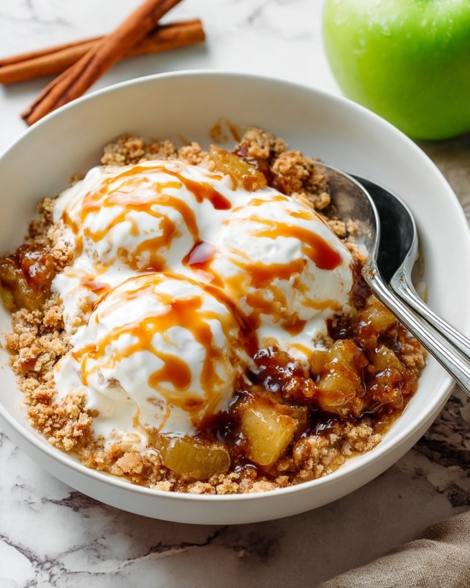 A white bowl holds a dessert with two large scoops of white creamy topping drizzled with orange-brown sauce. Below the cream is a crumbly, golden-brown textured layer mixed with soft chunks of cooked fruit that appear brownish-yellow and slightly shiny with syrup. The bowl is placed on a white marbled surface next to two metal spoons resting inside. In the background a green apple and cinnamon sticks are partially visible. Photo taken with an iphone --ar 4:5 --v 7