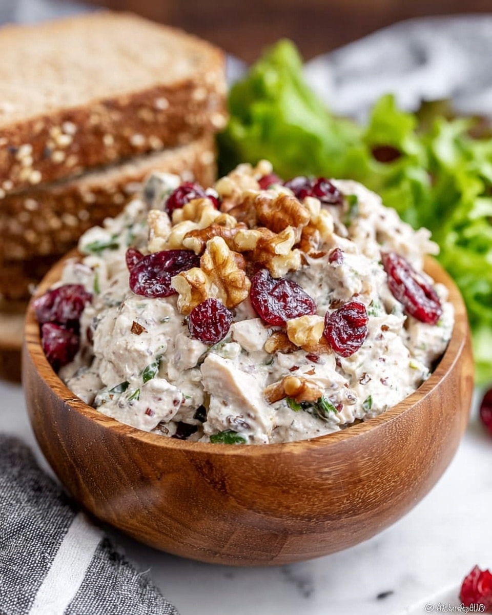 The image shows a wooden bowl filled with a creamy white chicken salad mixed with green herbs, small bits of walnuts, and dried red cranberries. On top of the salad, more walnuts and bright red cranberries are sprinkled, creating a colorful contrast. In the background, there are slices of whole grain bread with visible seeds stacked behind the bowl, and fresh green lettuce leaves slightly blurred. The bowl is set on a white marbled surface with a grey and white striped cloth partially visible near the base, giving a fresh and natural look to the scene. Photo taken with an iphone --ar 4:5 --v 7