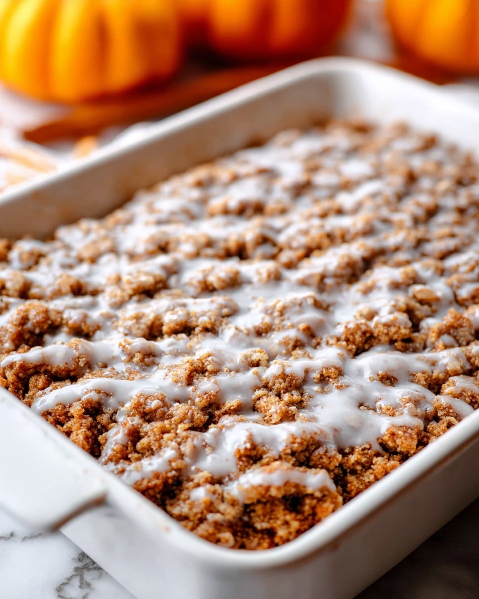A close-up view of a white rectangular deep baking dish filled with a crumble dessert. The top layer is rough and bumpy with a golden brown crumbly texture mixed with small clumps. Scattered unevenly over the crumble is a white icing glaze that looks smooth and slightly thick. The background shows a soft white marbled surface with blurred orange pumpkins adding a warm tone. Photo taken with an iphone --ar 4:5 --v 7