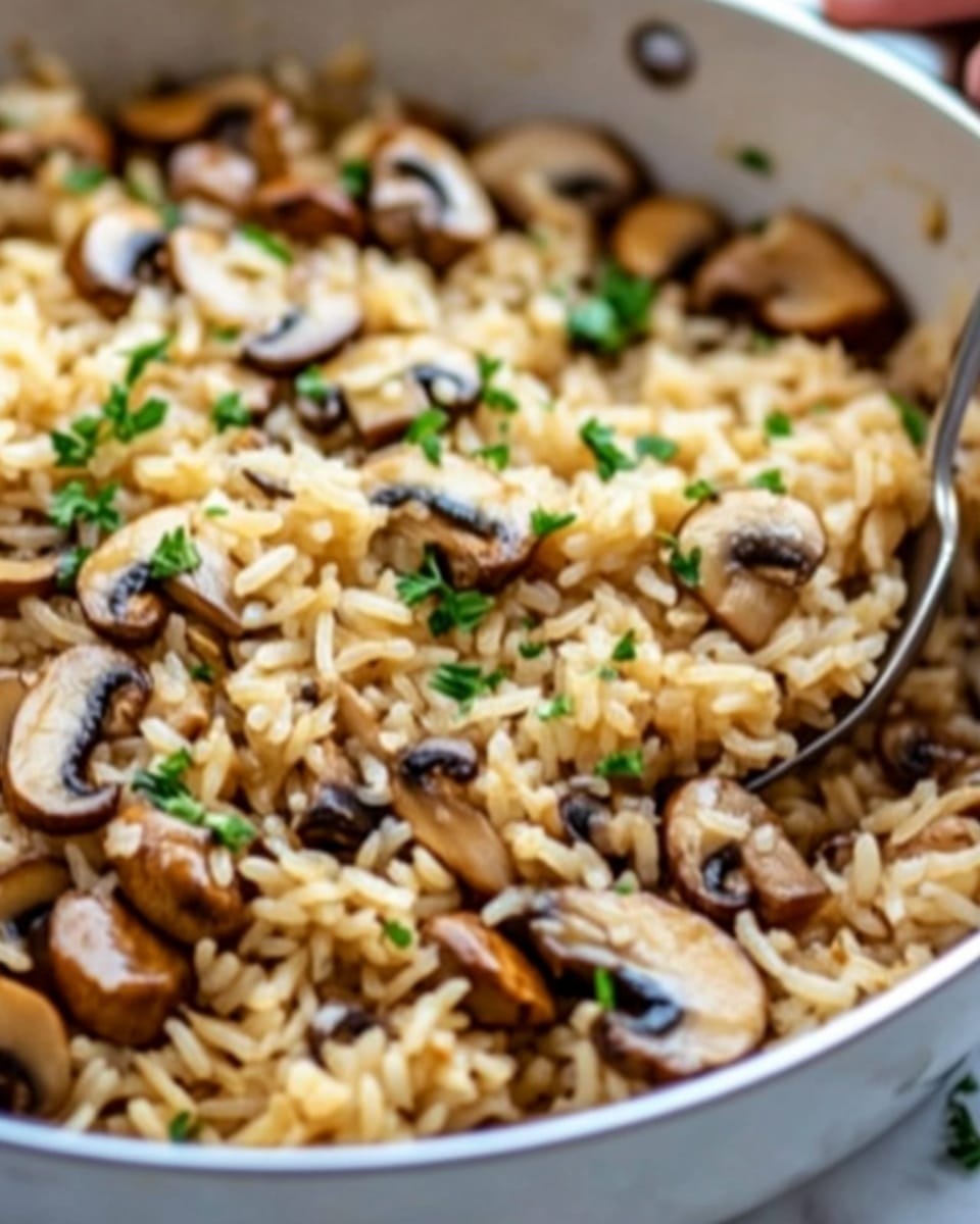 A close-up view of a white pan filled with cooked rice mixed with sliced brown mushrooms and small green herb pieces sprinkled on top. The rice looks soft and light brown, while the mushrooms are evenly spread throughout, showing a glossy texture. A shiny metal spoon rests inside the pan, partly buried in the rice, with a woman's hand holding the handle. The pan sits on a white marbled surface with soft natural light highlighting the colors. Photo taken with an iphone --ar 4:5 --v 6.1