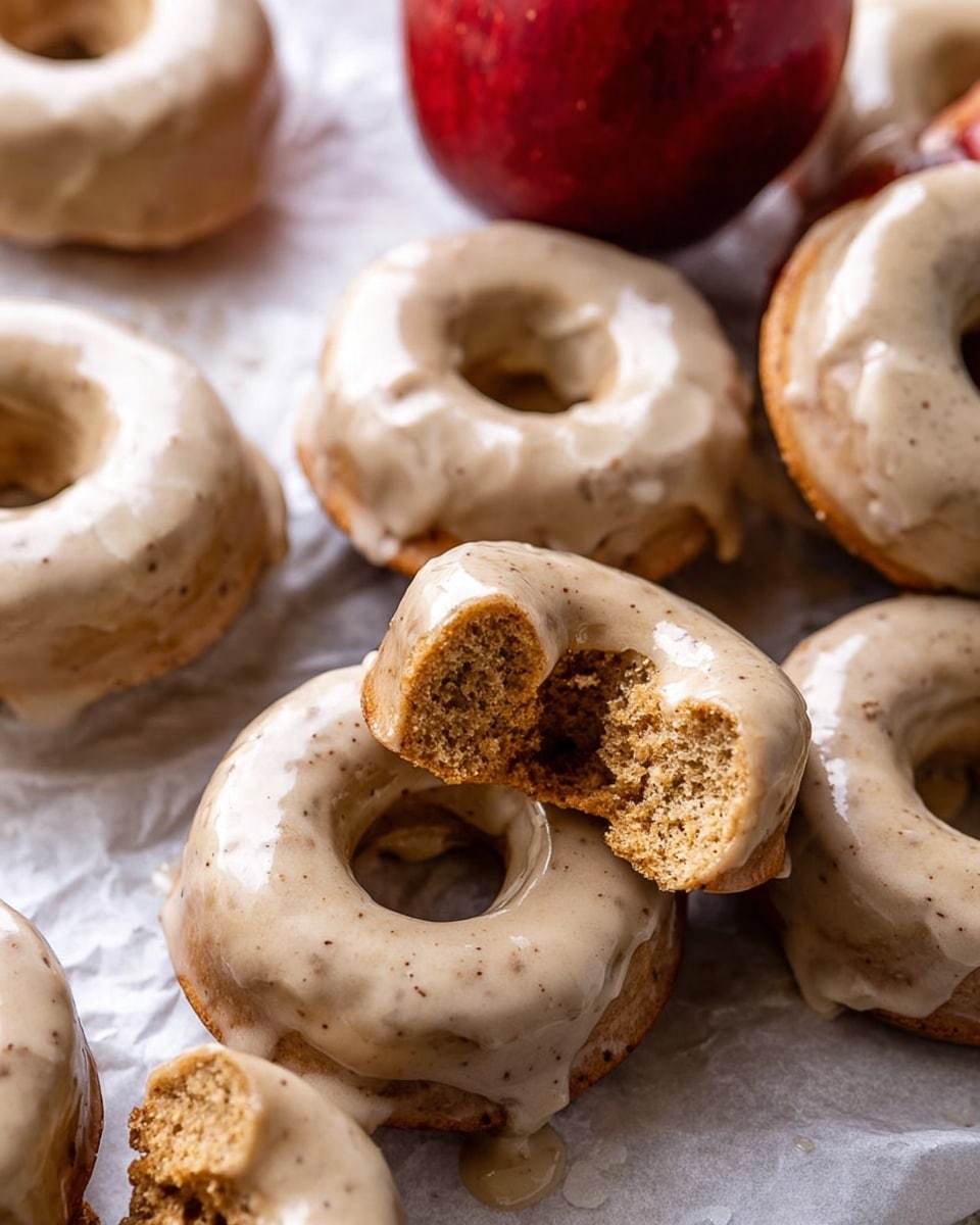A group of round donuts with smooth light beige frosting sit on crumpled white parchment paper on a white marbled surface. Some donuts are whole, showing their soft crumb inside with a golden brown color, while others are broken into pieces with crumbly texture. A shiny red apple with a green stem is nestled among the donuts, adding a bright contrast. There is also a white cup with a red handle filled with a light brown drink beside the donuts, and star anise pods are scattered around for decoration. photo taken with an iphone --ar 4:5 --v 7