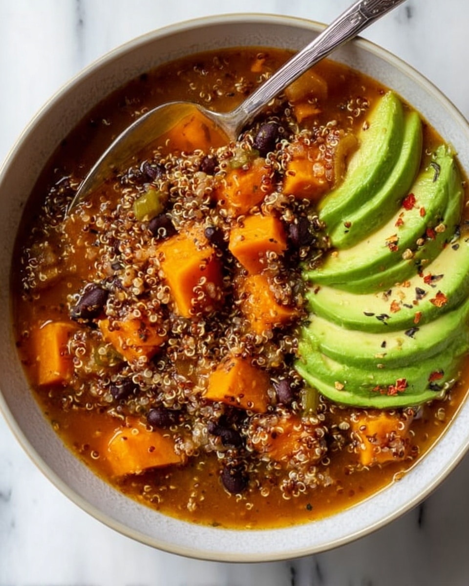 A white bowl filled with a thick soup made of small, light brown quinoa grains, black beans, and orange chunks of cooked sweet potato, all mixed in a reddish-orange broth. On one side of the bowl, there are thin slices of vibrant green avocado sprinkled with black pepper and small red flakes. A silver spoon rests inside the bowl, partially covered by the soup. The bowl sits on a white marbled surface. Photo taken with an iphone --ar 4:5 --v 7