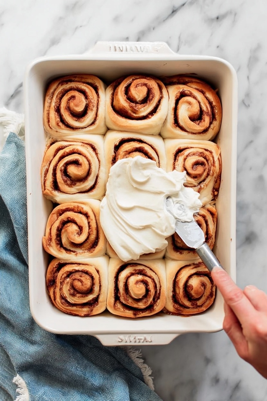 A white rectangular baking dish holds twelve cinnamon rolls arranged in a 3 by 4 grid. Each roll shows a spiral pattern with light brown dough and darker brown cinnamon filling. A woman's hand is spreading smooth white frosting over the center rolls with a silver spreading knife. The dish sits on a white marbled surface with a folded blue cloth nearby. Photo taken with an iphone --ar 2:3 --v 7 - Classic Homemade Cinnamon Rolls with Cinnamon Sugar Glaze, cinnamon roll recipe, easy cinnamon rolls, fluffy cinnamon rolls, homemade cinnamon roll glaze