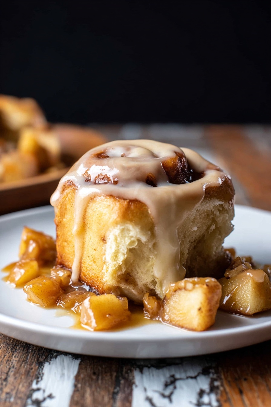 A cinnamon roll with visible swirls and a light golden brown color, topped with thick creamy icing that drapes over the edges, sitting on a white plate. Surrounding the roll are several torn pieces showing a soft, fluffy inside with a slightly darker cinnamon layer. Small chunks of a caramel-like sauce with bits of apple sit around the base, adding a shiny texture. The background is dark, and the plate rests on a rustic wooden surface with a white marbled texture. photo taken with an iphone --ar 2:3 --v 7 - Apple Cinnamon Rolls with Brown Butter Maple Icing, fall cinnamon roll recipe, apple cinnamon roll ideas, cozy weekend breakfast recipes, homemade cinnamon roll glaze