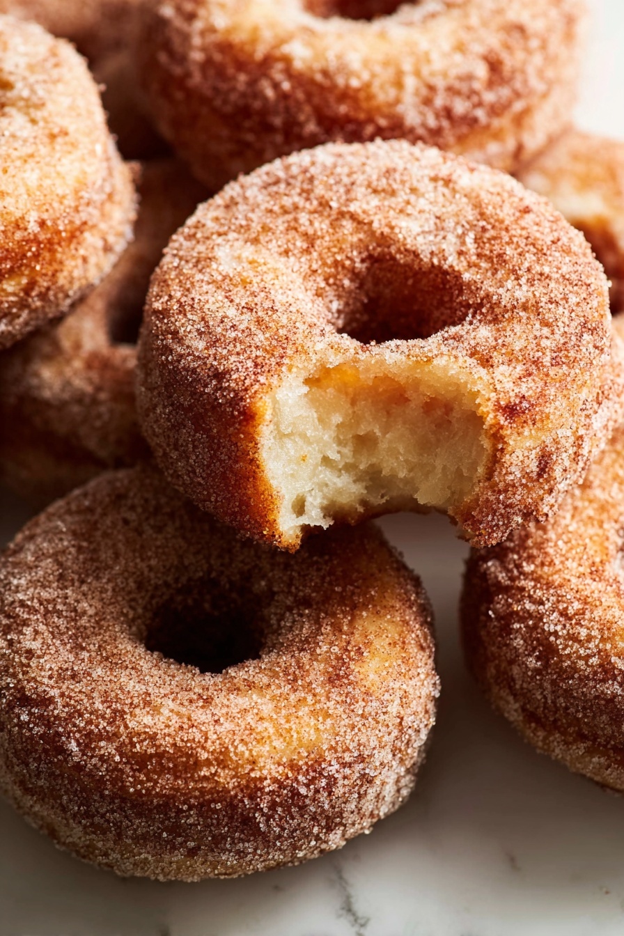 A stack of three sugar-covered donuts sits on a white marbled surface, with the top donut showing a bite taken out, revealing a soft, light brown inside. Each donut has a rough texture coated evenly in sugar, giving a slightly grainy look. The donuts are arranged one on top of the other, with the bitten one on the top, the middle and bottom donuts fully intact, all round with a visible center hole. The lighting softly highlights the sugar crystals and the golden brown color of the fried dough. photo taken with an iphone --ar 2:3 --v 7 - Apple Cider Donuts, fall apple cider donuts, homemade apple cider donuts, cozy spiced donuts, apple cider donut recipe