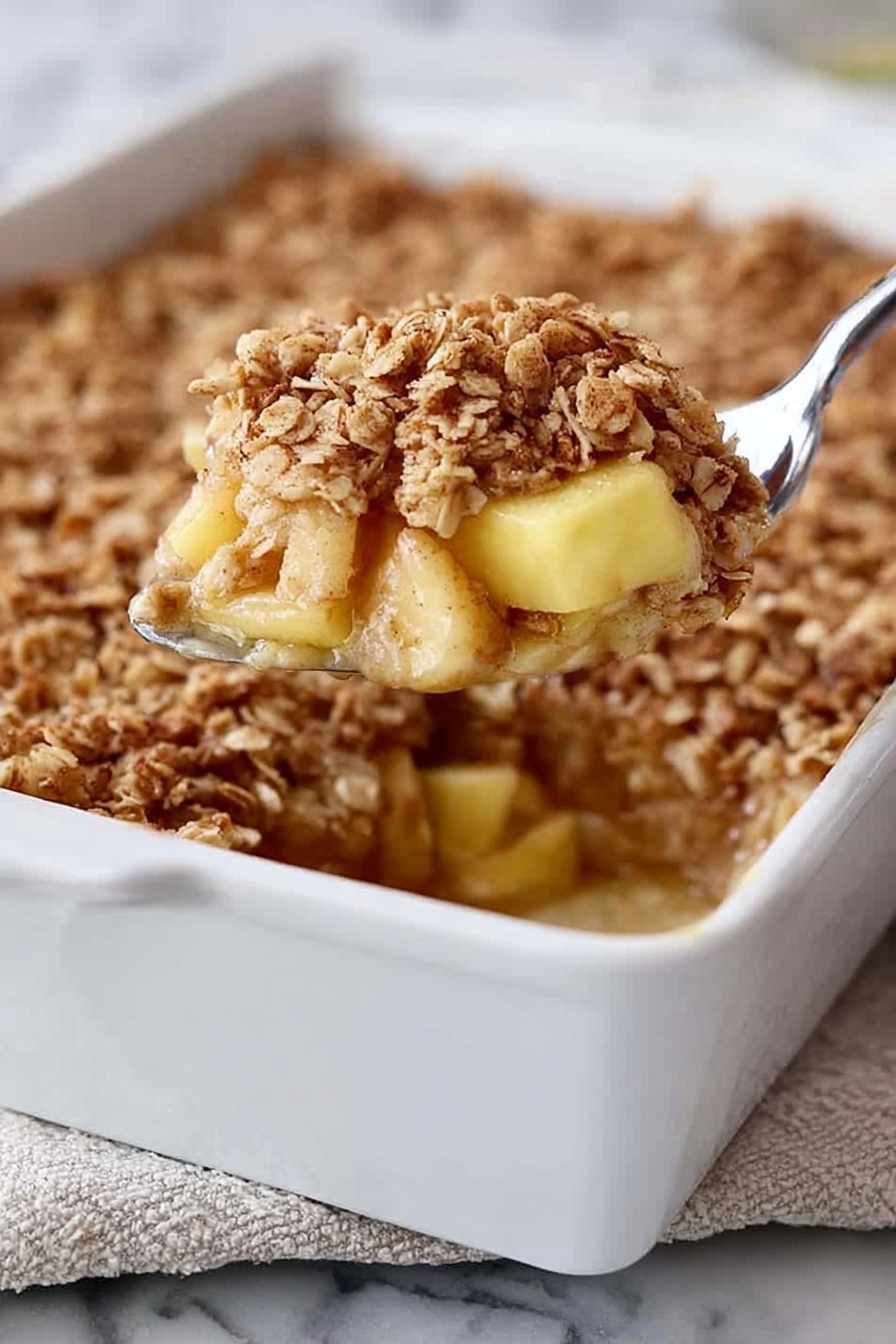 A white rectangular baking dish filled with a dessert that has three visible layers: at the bottom, soft light yellow chunks of apple, in the middle, a slightly sticky cinnamon-spiced syrup, and on top, a thick layer of golden brown oat crumble with a rough texture. A silver spoon is lifting a portion from the dish, showing the layers clearly. The dish is placed on a white marbled surface with a bit of a folded cloth underneath. Photo taken with an iphone --ar 2:3 --v 7 - Amish-Style Apple and Cinnamon Baked Oatmeal, healthy baked oatmeal with apples and cinnamon, comforting breakfast recipes, easy make-ahead breakfast ideas, cozy fruit oatmeal