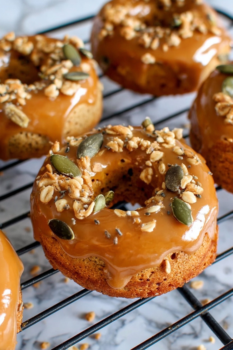 The image shows several small round donuts with a shiny caramel glaze on the top layer, sitting on a black cooling rack. The caramel glaze is smooth and glossy, covering the top and sides of the donuts. On top of the caramel layer, there are scattered crunchy toppings made of light brown crumbles and green pumpkin seeds, adding texture and color contrast. The donuts have a golden brown base with a soft, slightly rough surface. The background has a white marbled texture that frames the cooling rack and donuts softly. photo taken with an iphone --ar 2:3 --v 7 - Pumpkin Donuts with Bourbon Caramel Glaze, pumpkin spice donuts, fall donut recipes, bourbon caramel glaze, cozy autumn treats