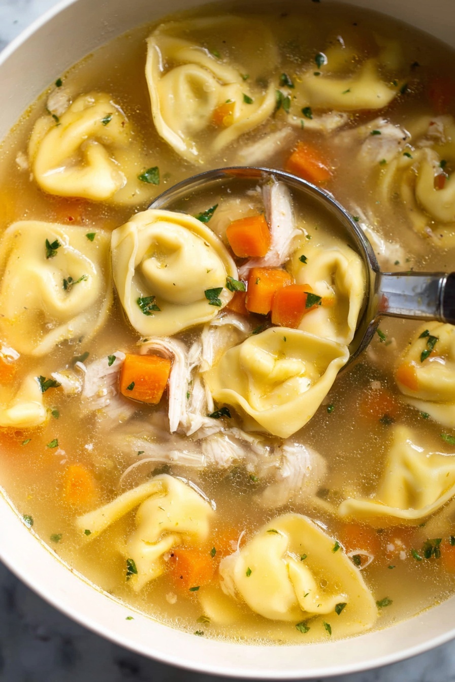 This image shows a close-up view of a bowl of clear broth soup filled with three main layers: the large, yellow tortellini pasta with a smooth, slightly wrinkled texture placed throughout; small, bright orange carrot cubes scattered evenly; and pieces of shredded white meat chicken mixed in. Small bits of green herbs float on top, adding a pop of color. A silver spoon with a long handle is scooping up the soup from the bowl, which is white, set on a white marbled surface. The broth has a light golden color and looks warm and inviting. Photo taken with an iphone --ar 2:3 --v 7 - Chicken Tortellini Soup, easy chicken tortellini soup, hearty tortellini soup, comforting chicken soup, quick weeknight soup