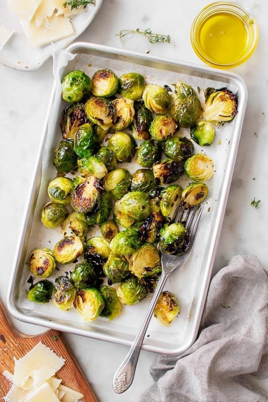 A white baking tray lined with white parchment paper holds a single layer of roasted Brussels sprouts, some halved and some whole, showing a mix of bright green and dark brown charred spots, giving a crispy texture. The tray is placed on a white marbled surface with a silver fork resting in the bottom right corner inside the tray. To the right, there is a small glass jar filled with yellow oil, and at the bottom left corner, part of a wooden board with thinly shaved white cheese is visible, along with a grey cloth nearby. The overall scene is bright and fresh, highlighting the crispy roasted Brussels sprouts. photo taken with an iphone --ar 2:3 --v 7 - Lemon Parmesan Roasted Brussels Sprouts, crispy Brussels sprouts with lemon and Parmesan, easy Brussels sprouts side dish, healthy roasted Brussels sprouts, flavorful Brussels sprouts recipe