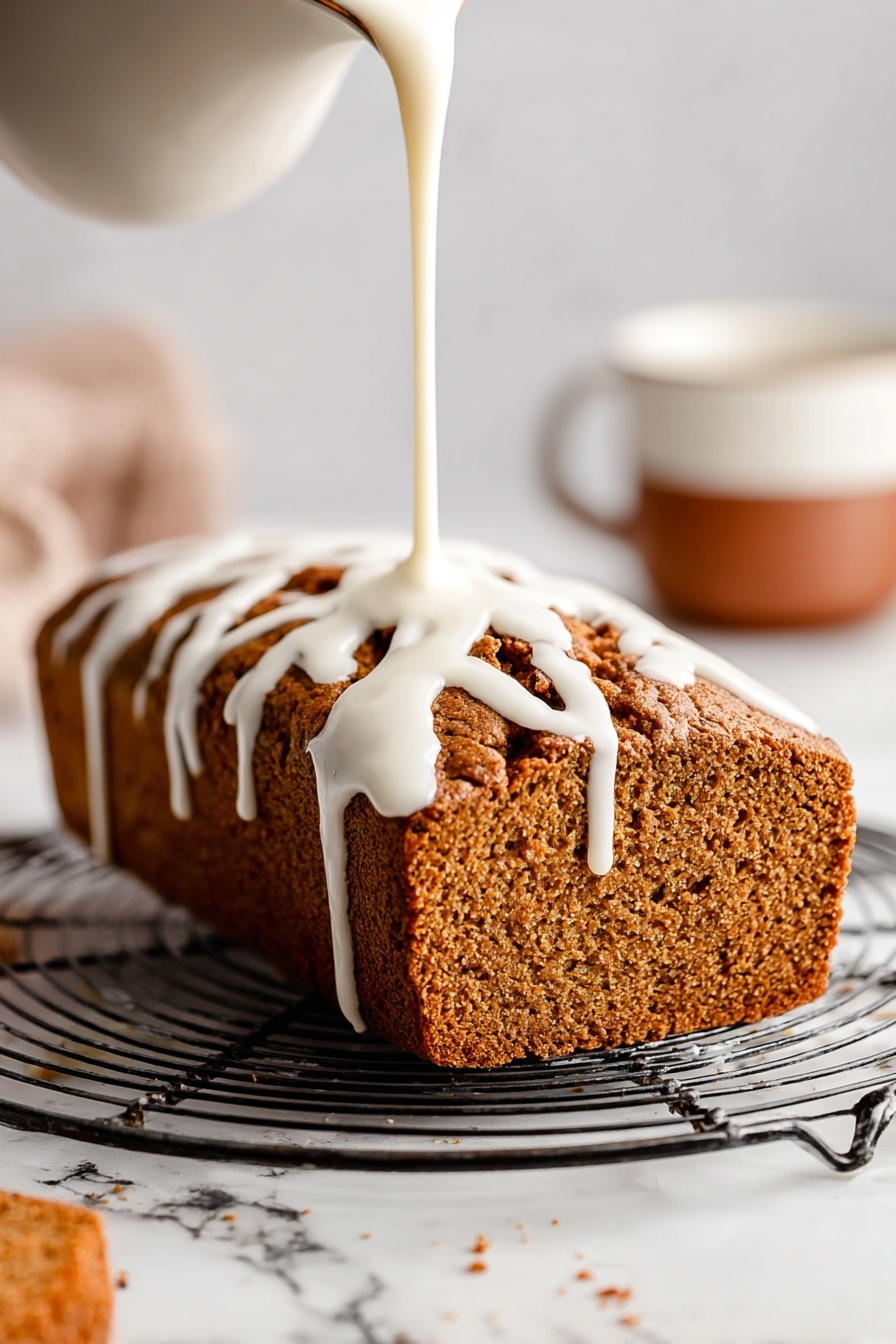 A loaf of brown textured bread sits on a round metal cooling rack with open grids. White icing is being poured over the top from above, flowing down the sides in thick, smooth ribbons. The loaf has a rough crust with deep cracks and a soft, porous interior visible on the side. In the background to the right, there is a blurred white cup with a brown lower half, all on a white marbled surface. Photo taken with an iphone --ar 2:3 --v 7 - Healthy Pumpkin Bread with Maple Glaze, pumpkin bread recipe, healthy pumpkin loaf, wholesome pumpkin bread, maple glaze pumpkin bread