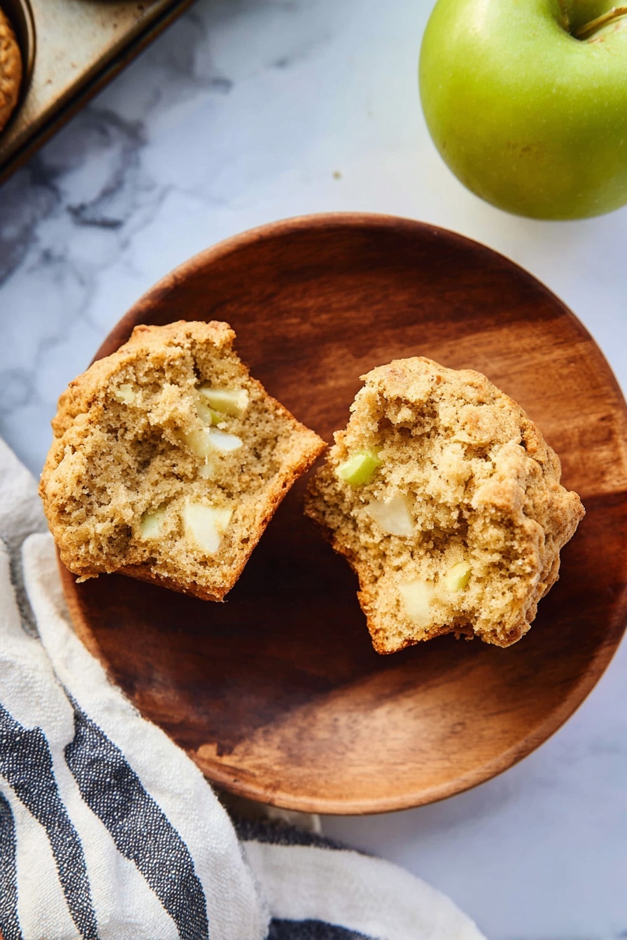 A round wooden plate on a white marbled surface holds a split muffin showing its soft, light brown crumb mixed with small white and slightly green apple pieces inside. To the right, a green apple sits partially in frame, and a striped white and dark blue cloth is partly visible beneath the plate and muffin tray corner. The muffin has a slightly golden crust with a rough texture. photo taken with an iphone --ar 2:3 --v 7 - Healthy Apple Muffins, healthy apple muffins recipe, wholesome apple muffin ideas, nutritious breakfast muffins, easy healthy muffin recipe