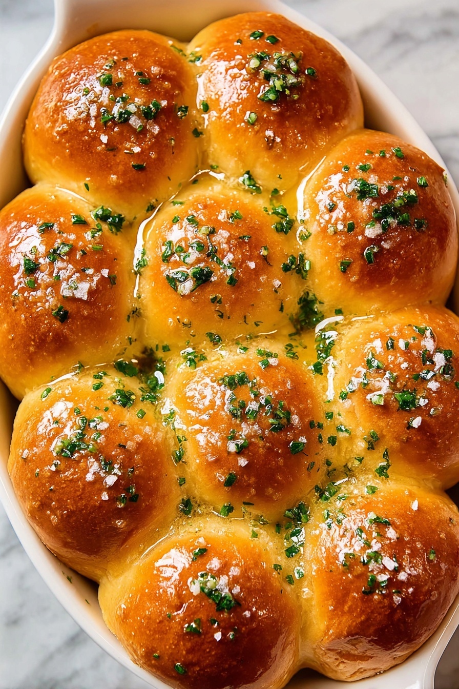 The image shows a white oval dish filled with 15 golden brown rolls arranged in a close cluster. Each roll has a smooth, shiny surface that looks soft and fluffy with a slight crisp on top. The rolls are topped with chopped green herbs spread evenly, adding fresh color contrast. There is a light glaze that makes the rolls appear moist, and small bits of coarse salt can be seen scattered on the surface. The dish is set on a white marbled texture background. photo taken with an iphone --ar 2:3 --v 7 - Garlic Butter Dinner Rolls, homemade bread rolls, fluffy dinner rolls, garlic bread rolls, soft bread recipes