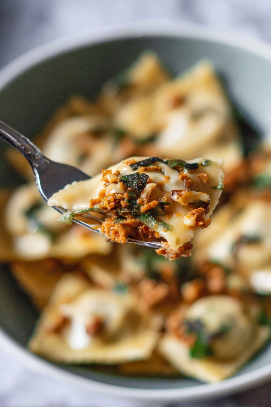 A close-up view shows a silver fork holding a piece of ravioli filled with a rich orange-brown meat mixture, topped with small dark green herb pieces and bits of light brown walnuts. In the blurred background, there is a white bowl filled with more ravioli, showing a mix of creamy beige pasta, white sauce, greens, and scattered walnut pieces, all set against a white marbled surface. The scene captures the soft texture of the ravioli and the fresh, rustic topping with a natural light that highlights the details. photo taken with an iphone --ar 2:3 --v 7 - Pumpkin Ravioli with Apple Sage Butter, pumpkin ravioli recipe, fall pasta dishes, homemade ravioli ideas, seasonal pumpkin pasta