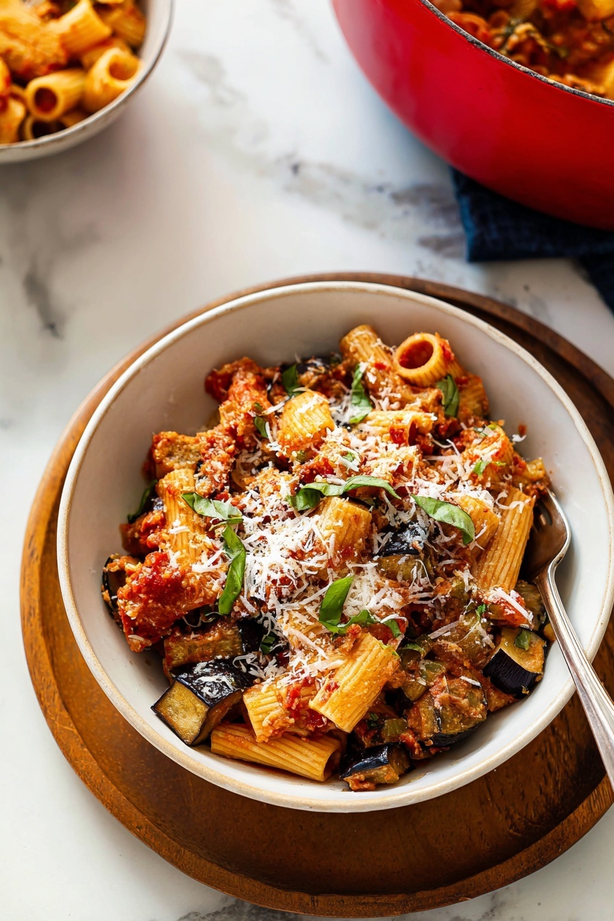 The image shows a white bowl placed on a round wooden tray on a white marbled surface. Inside the bowl, there are three main layers: the bottom layer has sautéed, golden-brown slices of eggplant, the middle layer consists of rigatoni pasta tubes mixed with a chunky red tomato sauce, and the top layer is sprinkled with shreds of white cheese and fresh green basil leaves. A silver fork is partially inserted into the pasta on the right side of the bowl. In the background and slightly to the left, a red pot filled with similar pasta can be seen. photo taken with an iphone --ar 2:3 --v 7 - Roasted Eggplant Pasta alla Norma, Sicilian eggplant pasta, eggplant pasta with basil and ricotta salata, hearty vegetarian Pasta alla Norma, easy Italian eggplant pasta