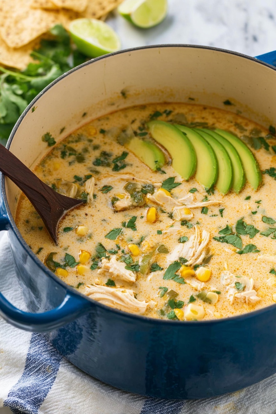 A large blue pot filled with creamy soup that has a light orange color with green cilantro leaves scattered on top. Inside the soup, there are chunks of white chicken, pieces of yellow corn, and bits of green vegetables. On one side, thin slices of light green avocado are placed in a neat row, partially submerged in the soup. A dark wooden spoon is stirring the soup from the left side. The pot sits on a white cloth with blue stripes on a white marbled surface. In the background, there are some light beige tortilla chips and lime wedges. Photo taken with an iphone --ar 2:3 --v 7 - Creamy White Chicken Chili, White Chicken Chili, Chicken Chili recipes, Easy White Chicken Chili, Hearty chicken soup