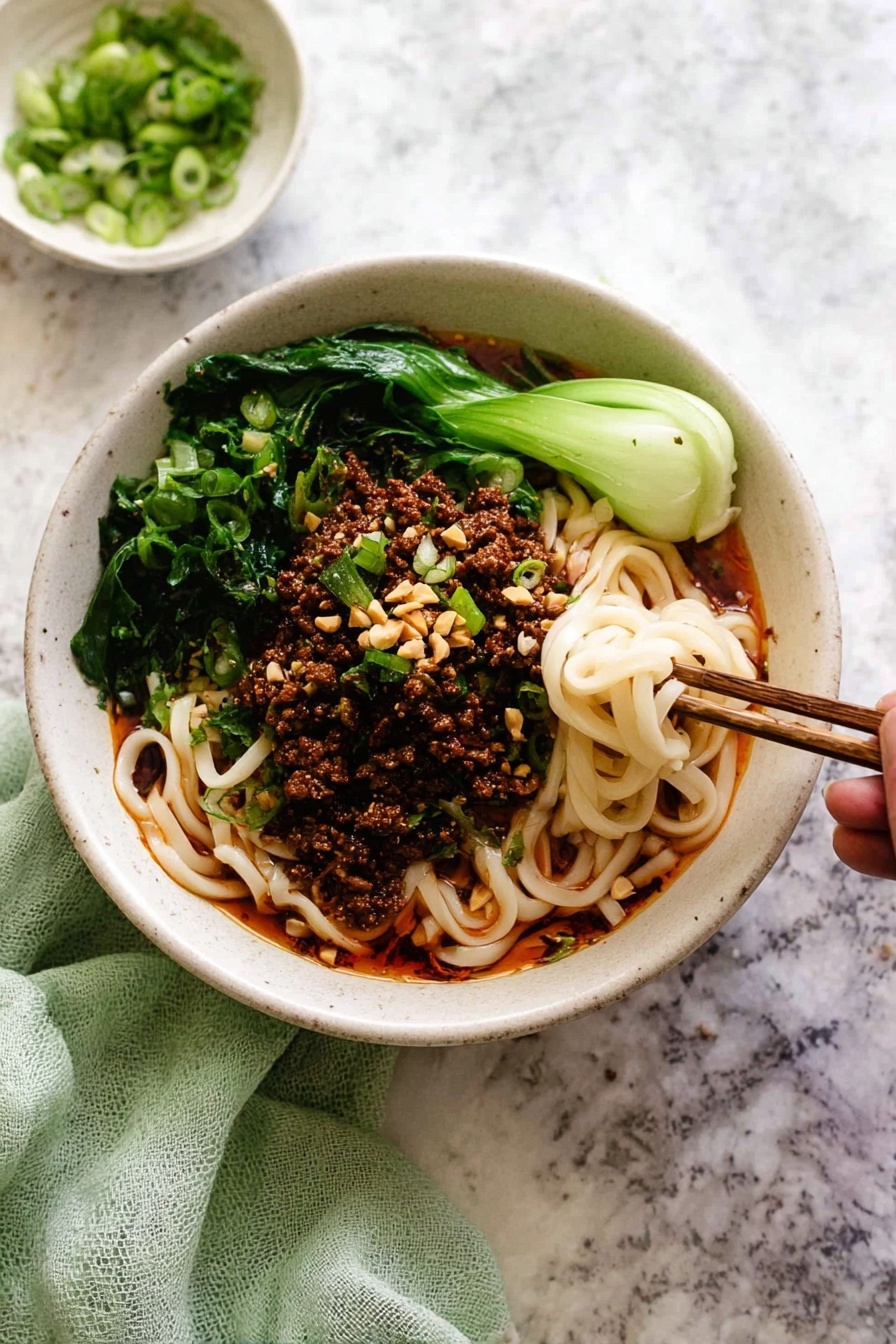 A white bowl filled with three main layers: at the bottom, thick white noodles mixed with a red oily sauce that gives a shiny texture; on top, a layer of dark brown minced meat scattered evenly; around the edges, bright green leafy vegetables adding fresh color; garnished with sliced light green scallions and small beige crushed nuts. A pair of dark brown chopsticks hold a small twisted bunch of noodles dipped in the sauce at the right side of the bowl. Some scallions and nut bits are scattered on the dark surface around the bowl. photo taken with an iphone --ar 2:3 --v 7 - Spicy Dan Dan Noodles with Pork, Dan Dan Noodles recipe, Sichuan pork noodles, spicy noodle bowl, quick Asian noodle dish