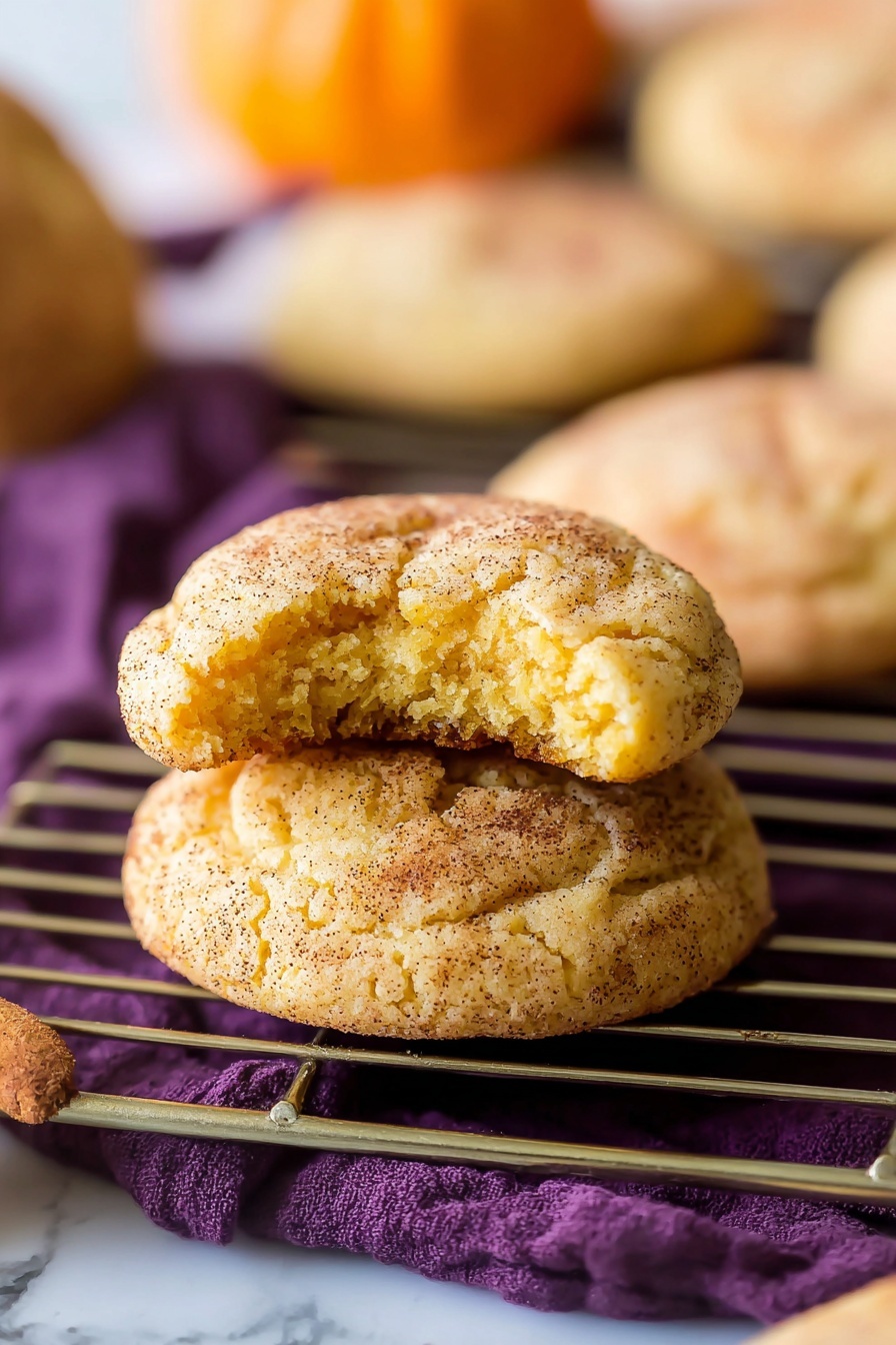 The image shows two soft, round cookies stacked on a silver cooling rack over a deep purple cloth, all set on a white marbled surface. The top cookie has a bite taken from it, revealing a light orange-yellow interior with a soft, cakey texture. Both cookies have a cracked surface speckled with dark cinnamon-like powder and a sandy sugary texture that makes them look warm and fresh. In the blurred background, there are more similar cookies and a small orange pumpkin visible. Photo taken with an iphone --ar 2:3 --v 7 - Pumpkin Snickerdoodle Cookies, fall pumpkin cookies, cinnamon sugar cookies, easy pumpkin cookie recipe, soft pumpkin cookies