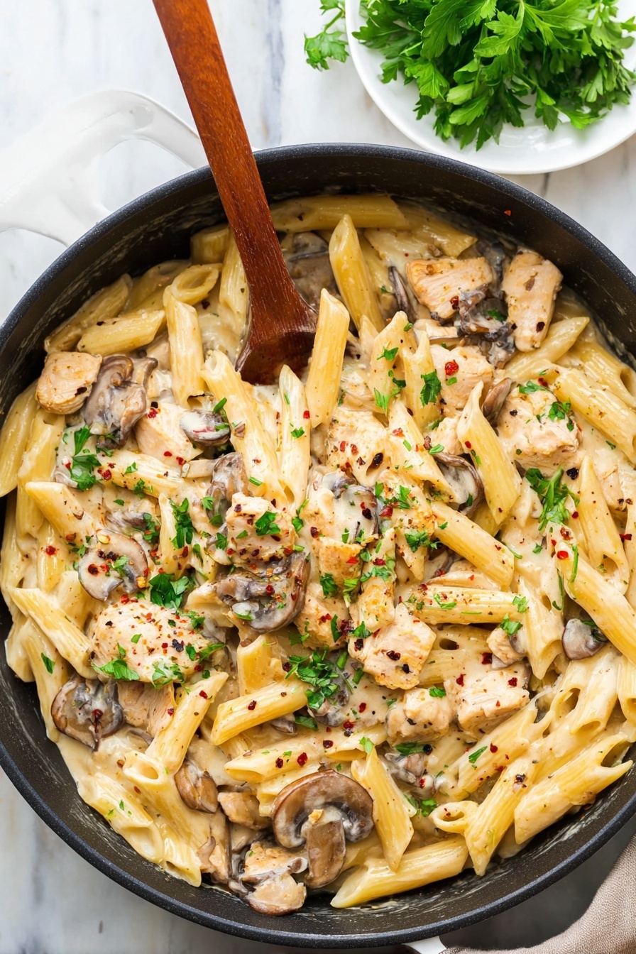 A black pan filled with creamy pasta made of three main layers: the base layer of pale yellow penne pasta, the middle layer of light brown cooked mushroom slices, and the top layer of small light beige pieces of chicken mixed in a creamy sauce. Green parsley leaves are scattered on top, adding bright green spots, with tiny black and red pepper flakes sprinkled over all. A wooden spoon rests inside the pan, partially covered by the pasta. The pan is placed on a white marbled surface, with a small white plate of fresh parsley leaves in the top corner. photo taken with an iphone --ar 2:3 --v 7 - Creamy Chicken Marsala Pasta, Chicken Marsala Pasta, easy chicken pasta dinner, creamy mushroom pasta, weeknight chicken recipes