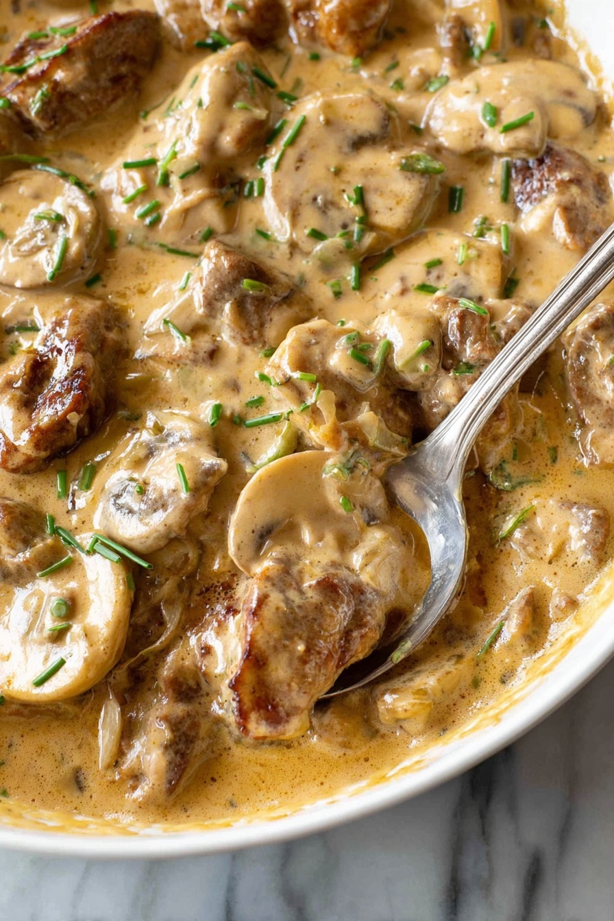 This image shows a close-up of a creamy dish with several layers visible. The base layer is a thick, light brown sauce with a smooth, slightly shiny texture, pooling around the other ingredients. Mixed in the sauce are slices of light beige mushrooms with soft, rounded edges and a slightly wet look. Also present are pieces of browned meat, which have a slightly rough texture and are coated in the same sauce. Small green chive pieces are scattered on top, adding little spots of bright green color throughout. A silver spoon is partially dipped into the dish, resting on the edge of a white bowl. The background is a white marbled surface. Photo taken with an iphone --ar 2:3 --v 7 - Quick Beef Stroganoff, easy beef stroganoff, creamy beef stroganoff, weeknight beef dinner, tender beef mushroom sauce