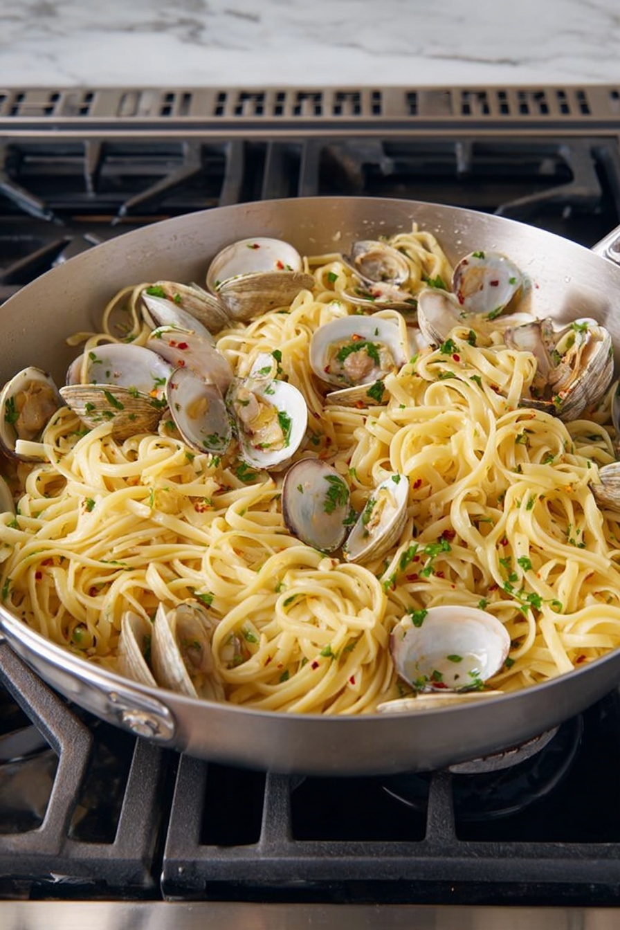 A large silver pan filled with cooked linguine pasta mixed with open clams distributed evenly throughout. The pasta is light yellow, soft and slightly shiny from sauce, with chopped green herbs sprinkled on top for color and freshness. Small bits of garlic and red chili flakes are visible mixed into the pasta, adding texture and flavor. The pan sits on a black stove with metal grates, surrounded by a white marbled surface. photo taken with an iphone --ar 2:3 --v 7 - Linguine with Fresh Clams and Lemon, seafood pasta with clams and lemon, easy seafood linguine, lemon pasta recipe, quick clam pasta dish