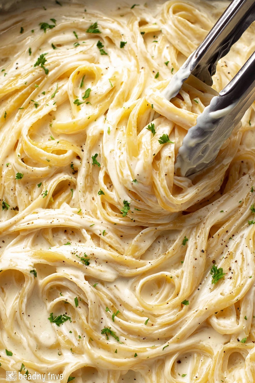 A close-up view of creamy fettuccine pasta in a thick white sauce filling the whole frame, with the pasta strands twisted and intertwined. The sauce is smooth and glossy with a light beige color and flecks of black pepper and small green parsley pieces sprinkled on top. Silver tongs are grabbing a bunch of pasta near the center, showing the rich texture of the sauce coating the noodles. The background is a white marbled texture. photo taken with an iphone --ar 2:3 --v 7 - Creamy Chicken Alfredo Pasta, Chicken Alfredo Recipe, Easy Alfredo Pasta, Homemade Chicken Alfredo, Restaurant-Style Alfredo