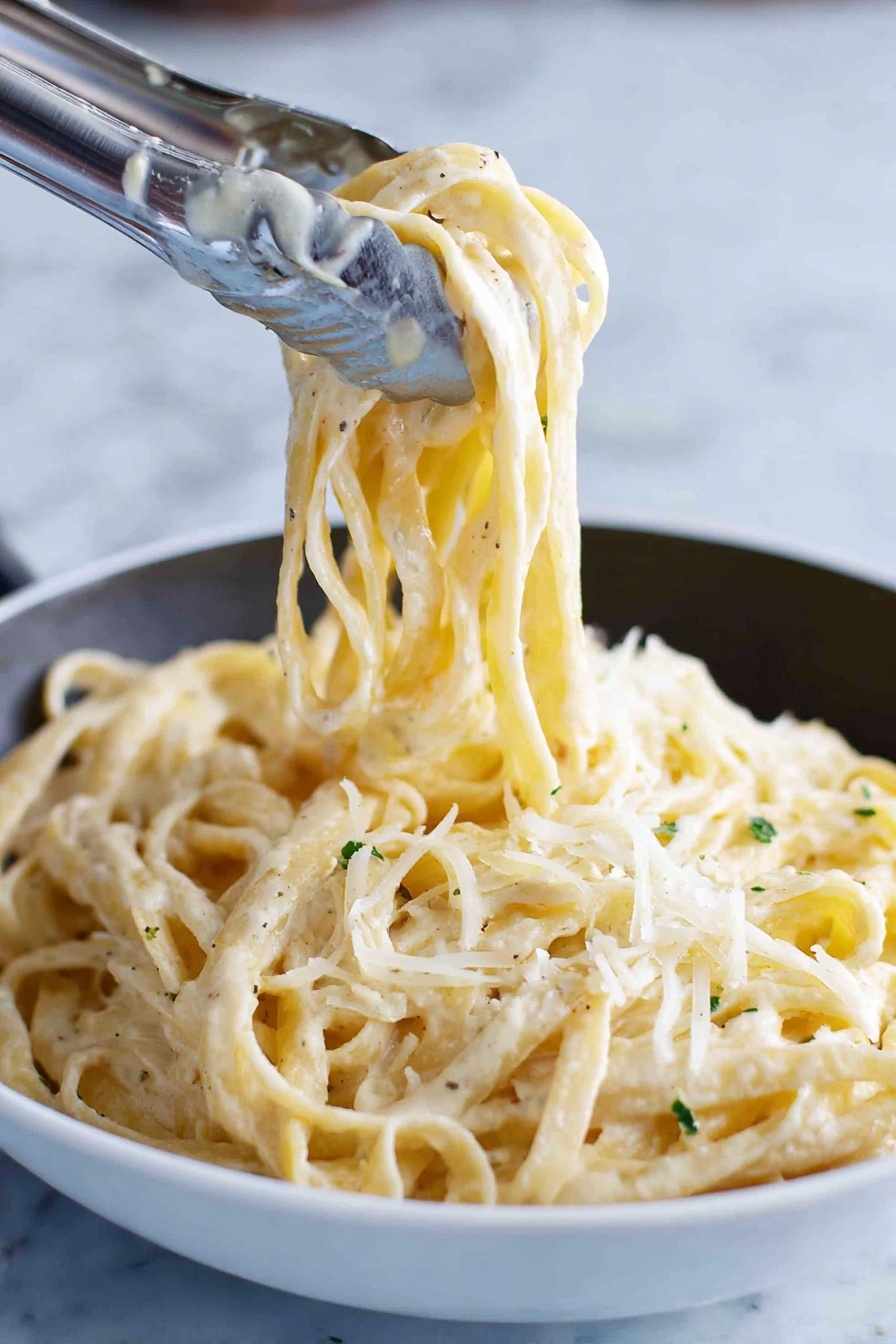 A close-up image shows thick noodles coated with a creamy white sauce, twirled and lifted by silver tongs above a white bowl filled with the same noodles. The noodles have a slightly glossy texture from the sauce and small green herb bits sprinkled throughout. Light shreds of pale cheese are scattered on top, adding a soft, fine texture contrast against the smooth noodles. The background is a white marbled surface. photo taken with an iphone --ar 2:3 --v 7 - Creamy Fettuccine Alfredo, authentic Italian pasta, homemade Alfredo sauce, easy pasta recipes, rich Italian fettuccine dinner
