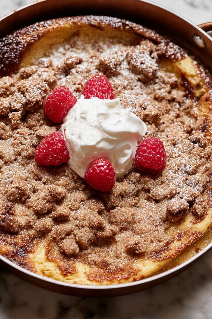 A round dessert with two main layers is shown in a pan. The bottom layer is a golden-brown, slightly risen cake with some darker browned spots. On top is a crumbly, light brown streusel layer that covers most of the surface unevenly. There is a dollop of white cream in the center with four bright red raspberries placed around and on it. A light dusting of powdered sugar is scattered over parts of the streusel and cake edges. The pan sits on a white marbled surface. photo taken with an iphone --ar 2:3 --v 7 - Brioche French Toast Casserole for Breakfast, easy holiday breakfast casserole, make-ahead French toast bake, rich breakfast casserole recipe, festive brunch ideas