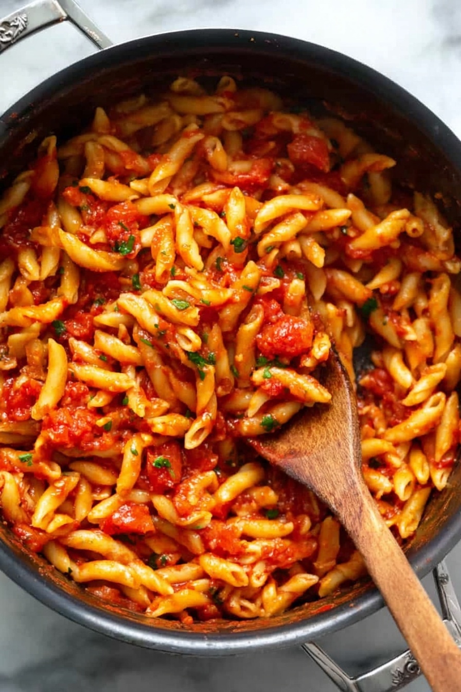 A black pot filled with a single layer of short, twisted pasta all covered evenly in a bright red tomato sauce with small bits of tomatoes and green herbs mixed in. A woman’s wooden spoon is stuck deep in the middle, lifting some pasta, showing the sauce clinging to the noodles. The pot is on a white marbled surface, the pasta looks soft and shiny with sauce. Photo taken with an iphone --ar 2:3 --v 7 - Spicy Penne Arrabbiata, Italian pasta with red pepper flakes, quick spicy pasta recipe, garlic tomato pasta, easy Penne Arrabbiata
