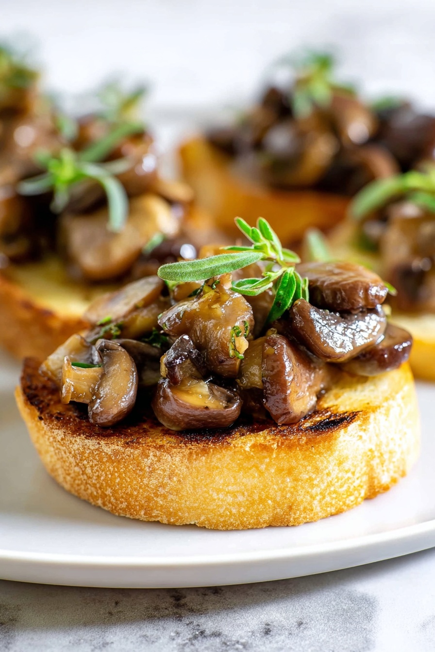 The image shows a close-up of a small round toast slice as the base, golden brown with light grill marks and a slightly rough texture. On top of the toast is a generous layer of sautéed mushrooms in varying shades of brown, some shiny from oil, with a mix of small and large mushroom pieces creating a textured and chunky look. The mushrooms are garnished with small fresh green herb sprigs placed on top, adding a pop of color. The toast with mushrooms is placed on a white plate set against a white marbled background. photo taken with an iphone --ar 2:3 --v 7 - Mushroom Thyme Crostini, mushroom crostini appetizer, easy mushroom crostini, savory mushroom crostini, mushroom and thyme bruschetta