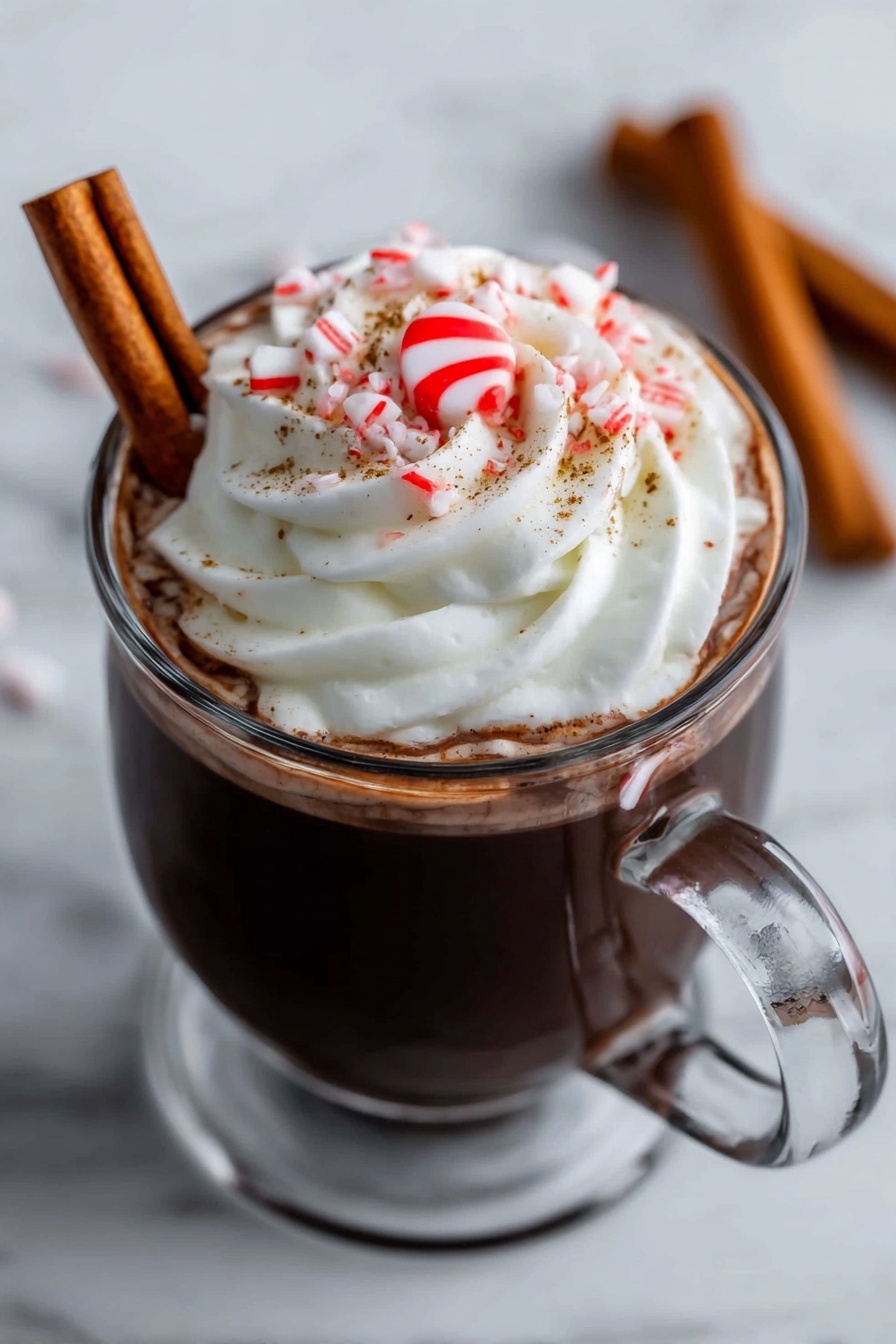 A clear glass mug filled with dark hot chocolate topped with a large swirl of white whipped cream. On top of the cream is a small red and white peppermint candy in the center, surrounded by some small pieces of crushed candy cane and fine brown sprinkles. A brown cinnamon stick is resting against the side inside the mug. The mug is placed on a white marbled surface. In the background, two more cinnamon sticks lie on the surface out of focus. A woman's hand is holding the handle of the mug. photo taken with an iphone --ar 2:3 --v 7 - Festive Peppermint Hot Chocolate, peppermint hot chocolate recipe, holiday hot chocolate, cozy winter drinks, creamy peppermint hot chocolate