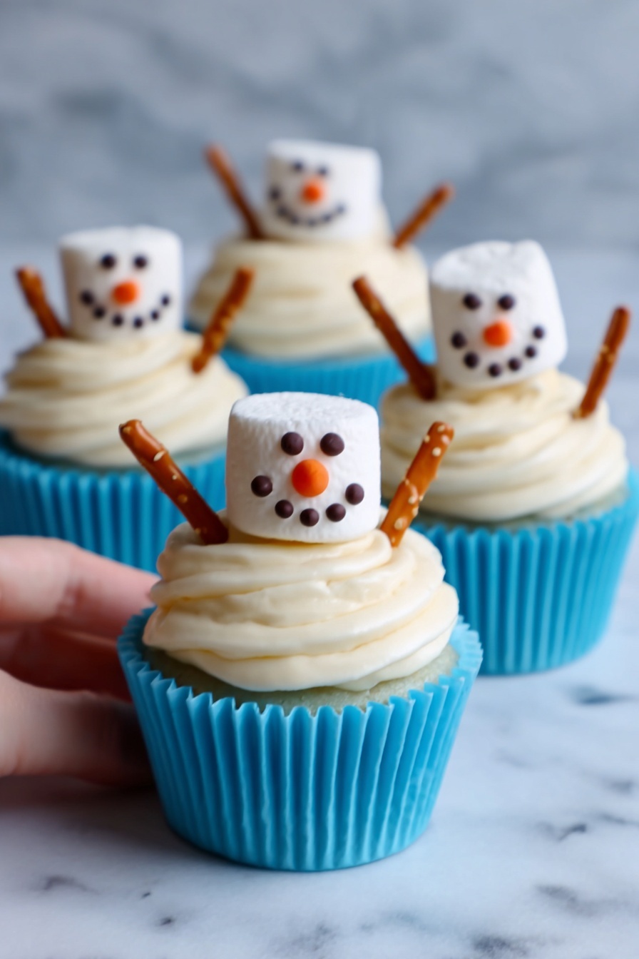 The image shows three cupcakes in white cupcake liners with bright blue bases, each topped with thick, creamy white frosting swirled in layers. On top of each cupcake sits a large marshmallow decorated like a snowman face with small dark chocolate or icing dots for eyes and mouth, and an orange triangular nose. Two pretzel sticks are inserted on opposite sides of the frosting, looking like the snowman's arms. The cupcakes are placed on a white marbled surface, with one woman's hand gently holding the front cupcake. The scene has bright, soft lighting. photo taken with an iphone --ar 2:3 --v 7 - Snowman Cupcake Muffins, winter cupcake decorations, adorable holiday muffins, chocolate muffin recipes, festive winter desserts