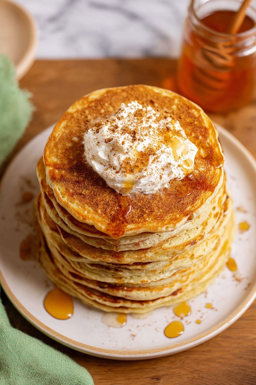 A stack of seven golden brown pancakes sits centered on a white plate with a subtle rim design, each pancake thick and fluffy with a smooth texture. On top of the stack is a generous dollop of cream-colored butter sprinkled with brown cinnamon powder, melting slightly with amber syrup drizzled over the pancakes in thin, uneven lines. The plate rests on a wooden table with a translucent glass jar of honey nearby, and a light green cloth is partially visible on the left side. The background features a white marbled texture. photo taken with an iphone --ar 2:3 --v 7 - Cinnamon Honey Butter, cinnamon honey butter spread, easy cinnamon butter, homemade cinnamon butter, versatile breakfast spread